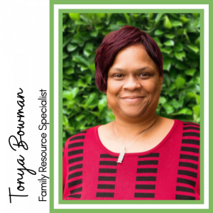 Image of smiling Black woman with short red hair. She is wearing a red and black patterned shirt standing in front of green leaves.