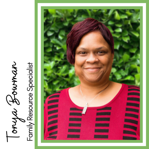 Image of smiling Black woman with short red hair. She is wearing a red and black patterned shirt standing in front of green leaves.